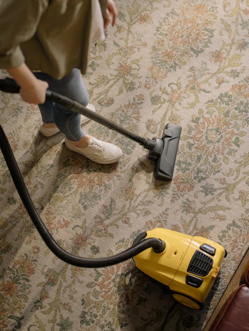 A person using a vacuum cleaner with a long hose on a patterned area rug in a domestic living space. The rug features floral motifs in muted tones and covers a wooden floor. The individual, dressed in