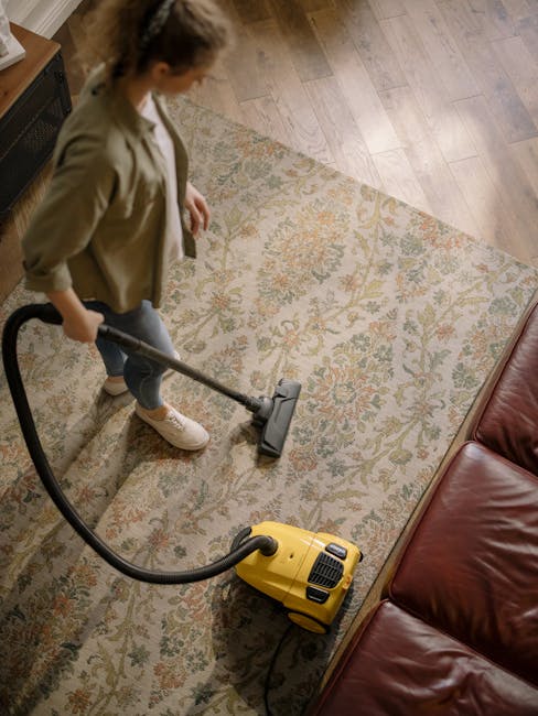 A person using a vacuum cleaner with a long hose on a patterned area rug in a domestic living space. The rug features floral motifs in muted tones and covers a wooden floor. The individual, dressed in